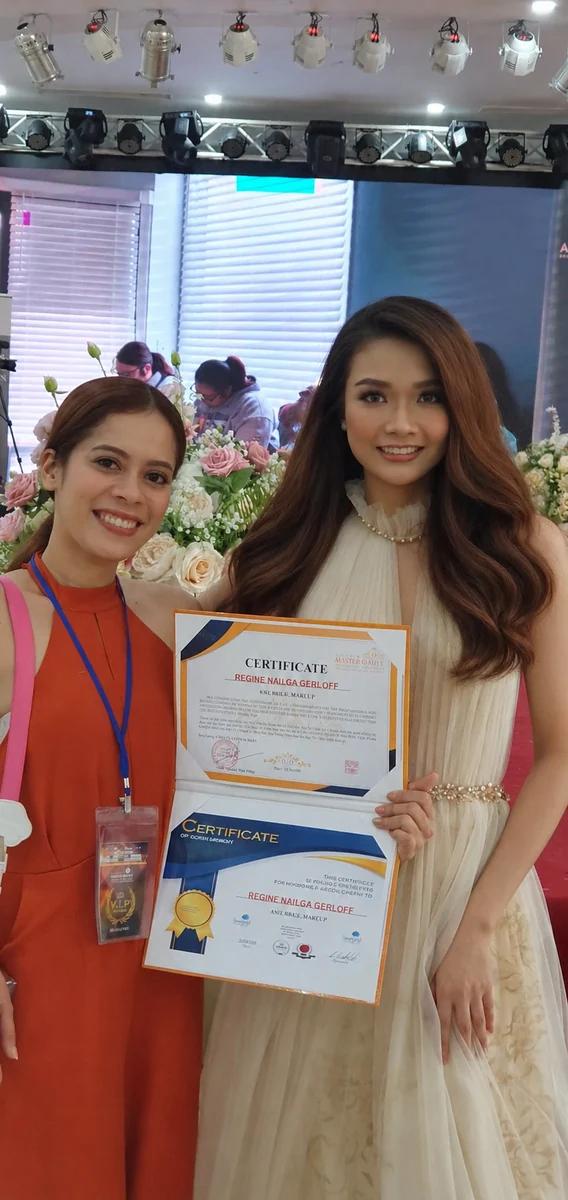 Two women smiling at a beauty event and holding open certificates, with flowers and stage lighting behind them