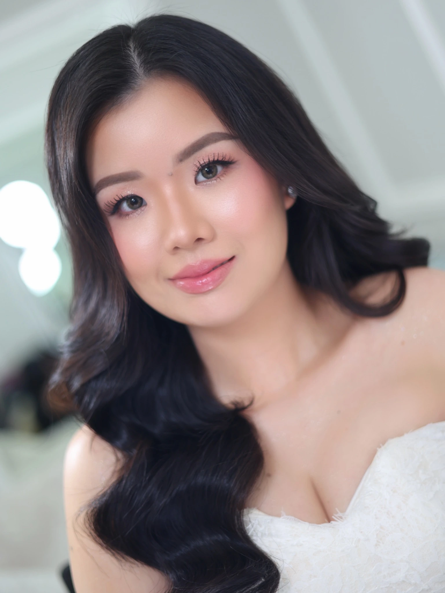 Close-up bridal portrait with long dark waves, softly shimmering eyes, pink blush and glossy lips, wearing a white off-the-shoulder gown