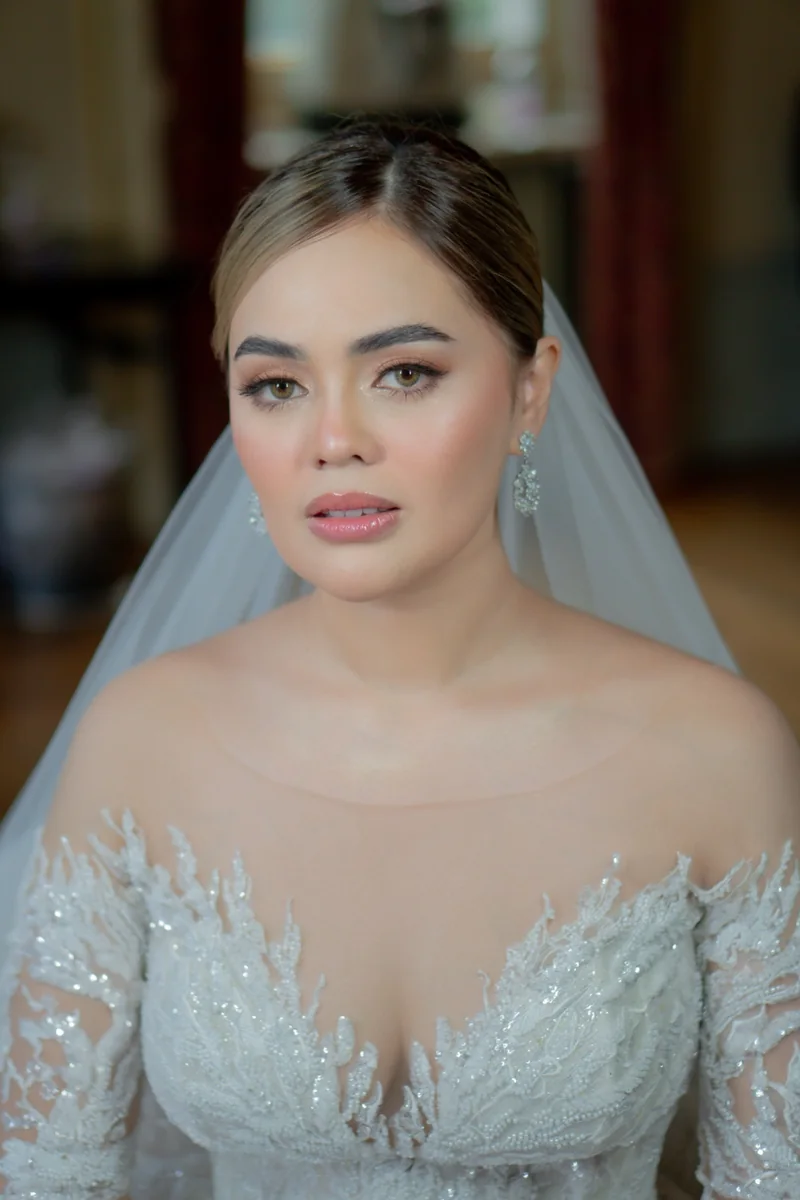 Close-up of a bride with softly defined eyes, rosy cheeks and glossy pink lips, wearing a veil and a beaded white gown