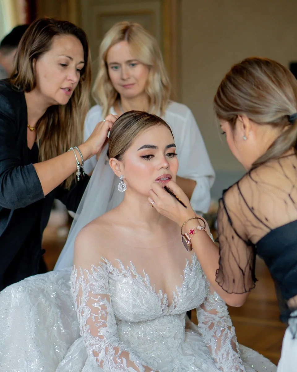 Makeup and hair preparation around a seated bride in a white gown, with a lip brush being applied