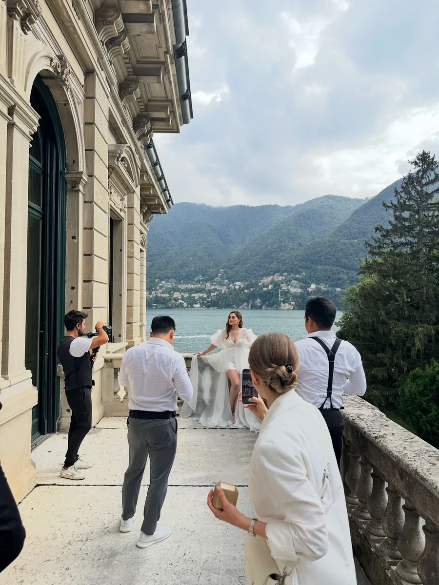 A photography crew capturing a woman in a white dress beside an ornate building overlooking a lake and wooded hills