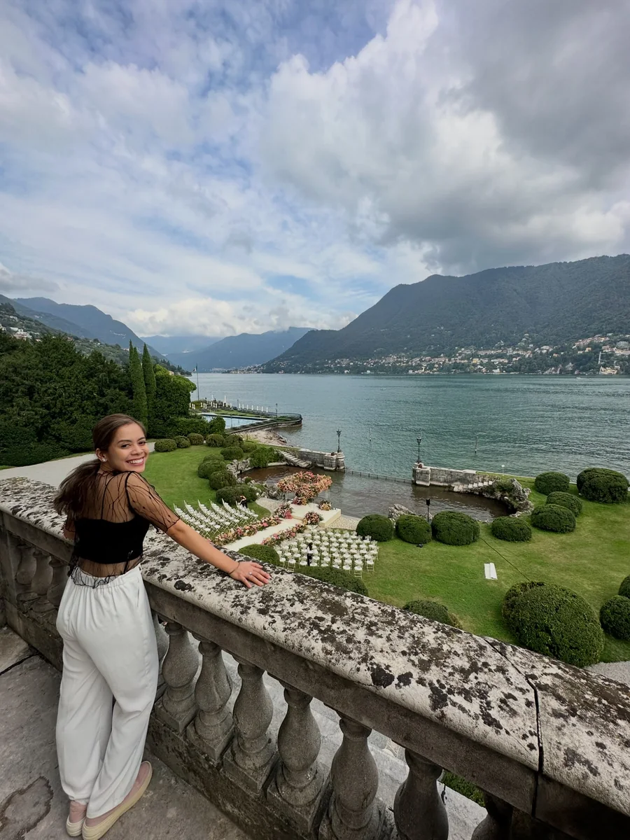 Smiling woman leaning against a stone terrace railing overlooking a lake, garden and outdoor wedding seating