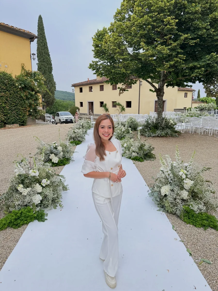 Smiling woman in a white outfit standing on a flower-lined outdoor ceremony aisle with white chairs and trees