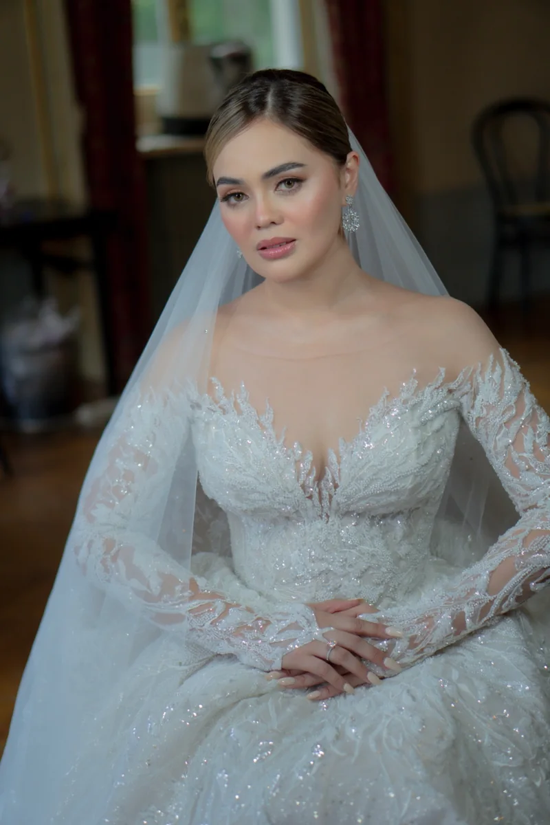 Seated bride with a veil and crystal earrings, wearing a detailed white gown and resting her hands in her lap