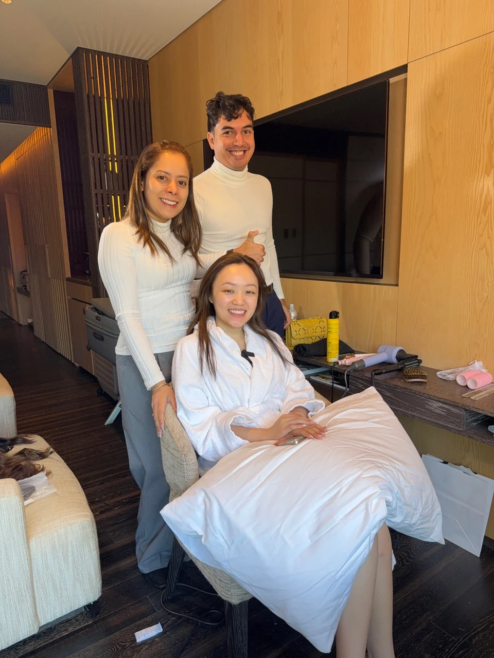 Bride seated in a white robe with two people standing behind her in a preparation room with hair styling tools