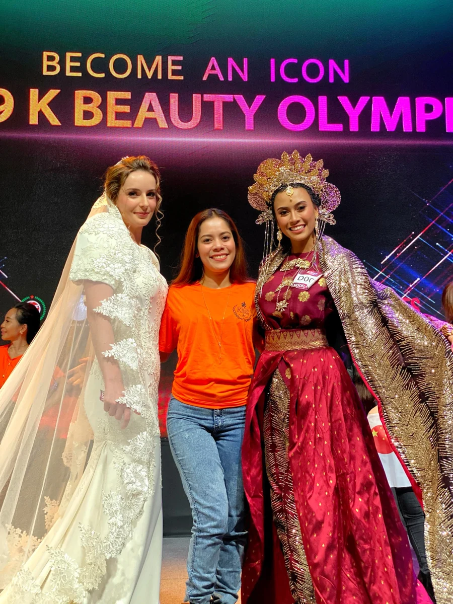 A participant in an orange shirt posing between a model in a lace bridal gown and a model in an ornate red-and-gold costume beneath K-Beauty Olympic signage