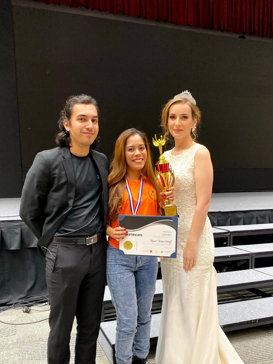 Three people posing by a stage, with a participant wearing an orange shirt and holding a certificate, medal and tall gold-and-red trophy beside a model in a lace gown