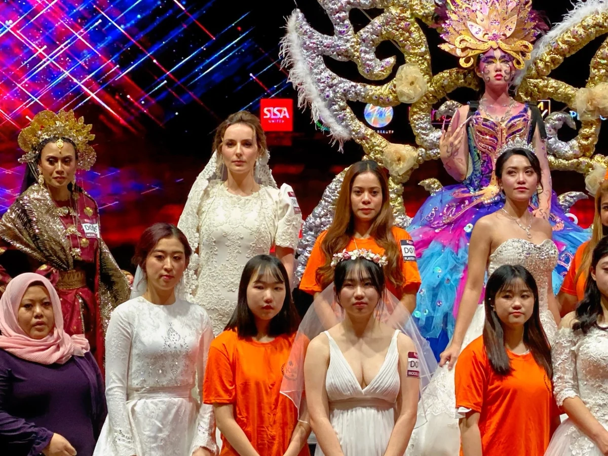 Competition participants and models posing on a stage with white bridal dresses and elaborate fantasy costumes