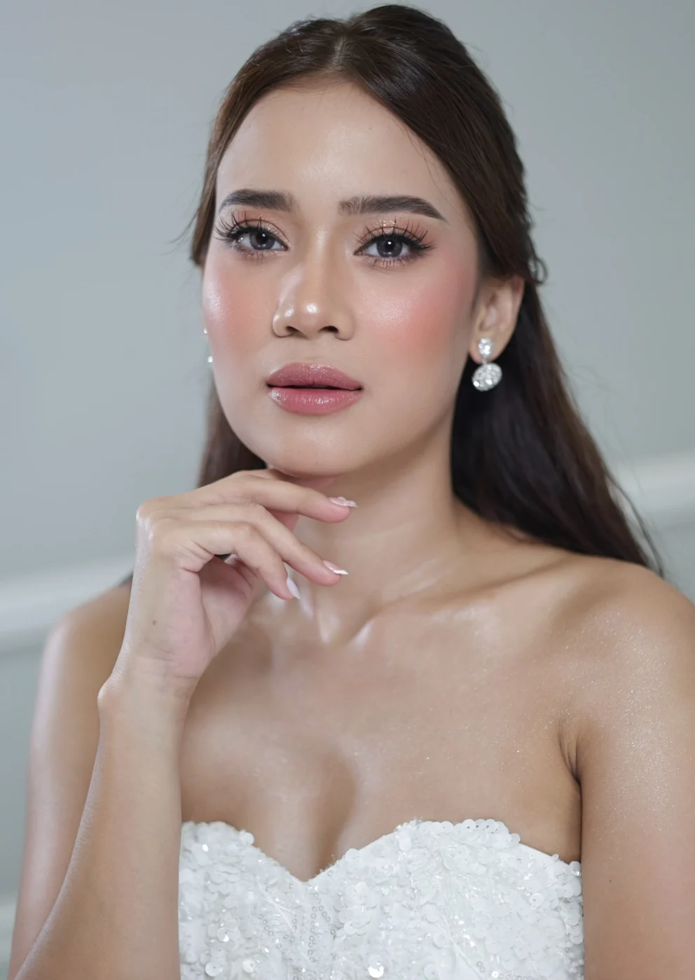 Portrait with a hand under the chin, rosy cheeks, soft pink lips, pearl-drop earrings and a strapless white gown