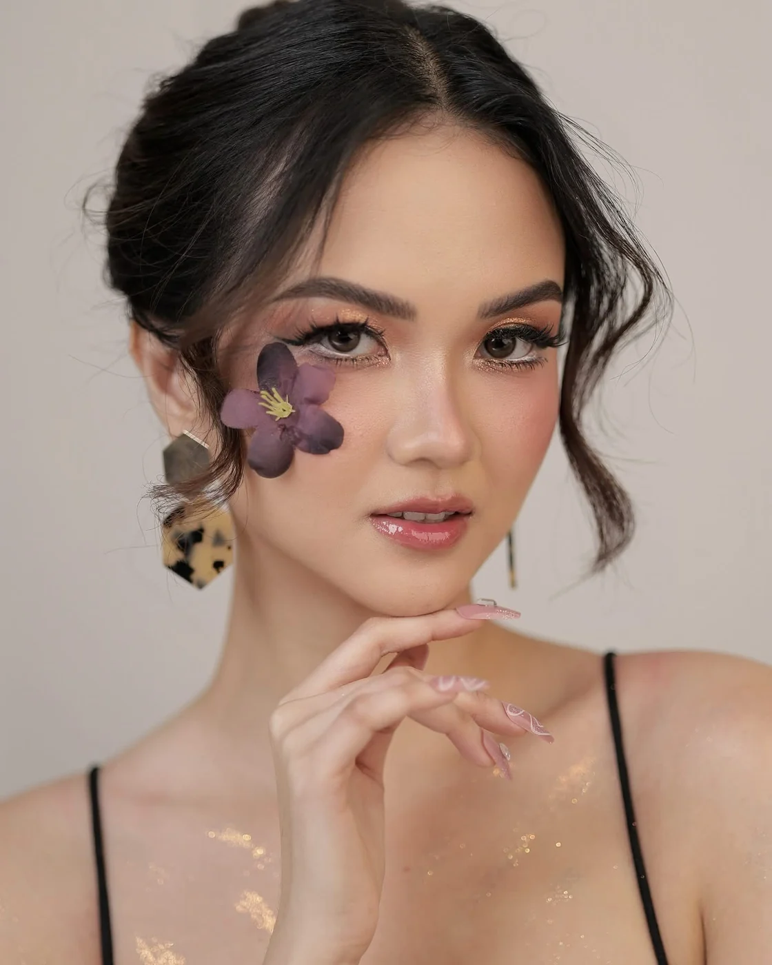 Beauty portrait with a loose updo, a purple flower beside one eye, a hand under the chin and soft nude-pink lips