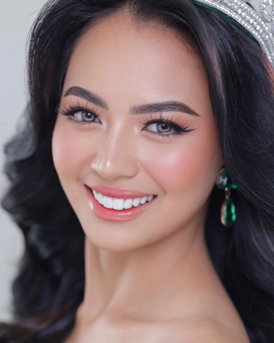 Smiling portrait with long dark hair, a jeweled tiara, green drop earrings and rosy pink makeup
