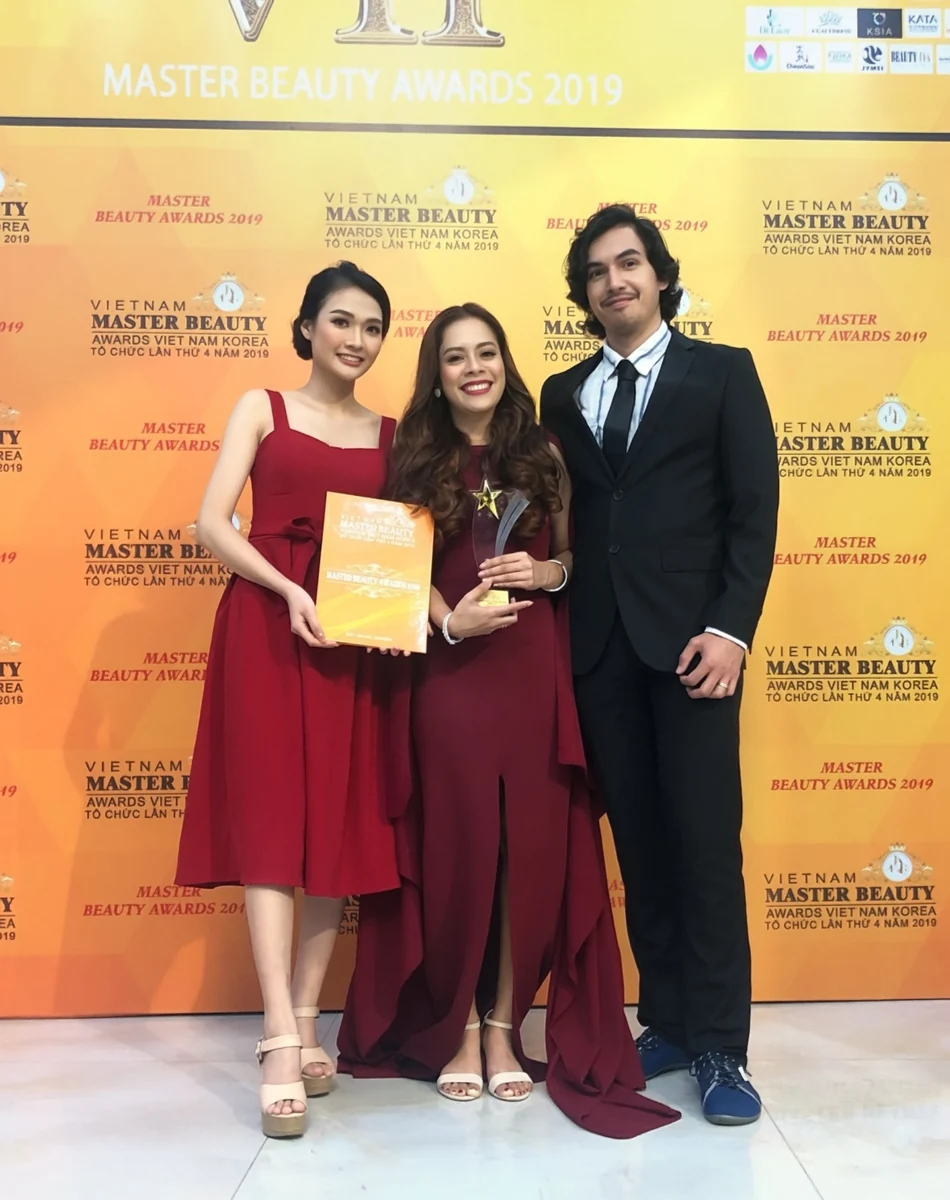 Three people posing at a Vietnam Master Beauty Awards 2019 backdrop, with a star-shaped trophy and an event certificate
